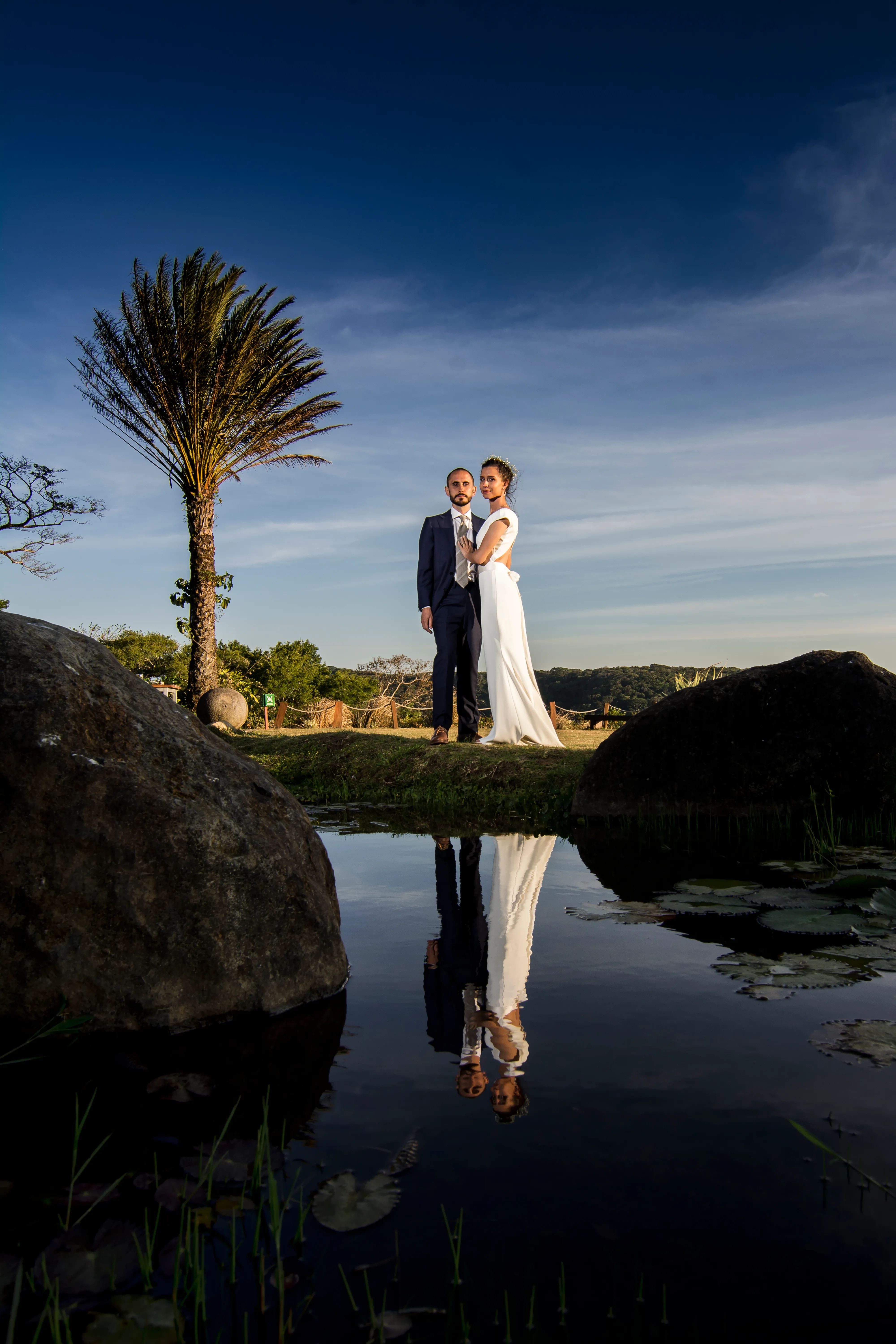 The bride and groom pose near our pond on the front lawn of our property. The sky is a deep blue with an ombre of clouds and the forest in the background.